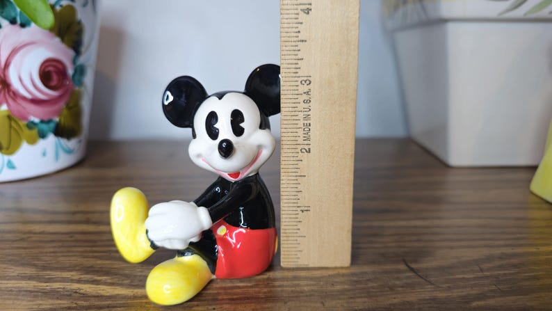 K&ouml;nnte beinhalten: Eine Keramikfigur von Mickey Mouse mit schwarzen Ohren, einem schwarzen K&ouml;rper, roten Shorts und gelben Schuhen. Die Figur ist etwa 7,6 cm hoch, mit wei&szlig;en Handschuhen und einem l&auml;chelnden Gesicht. Ein Holzlineal befindet sich daneben.