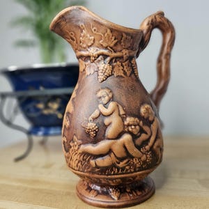 May include: A brown ceramic pitcher with a vine and grape design. The pitcher features a raised relief of three figures, two of which are nude.