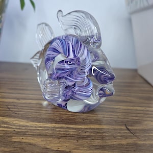 May include: A clear glass sculpture of a fish with purple and white swirls. The fish has fins and a detailed design. The sculpture is on a wooden surface, with a floral decorated pot and a white box in the background.