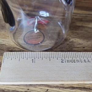 May include: A clear glass cup with a black handle is positioned near a wooden ruler. The ruler, marked in inches, is labeled "MADE IN U.S.A." The cup has a small, round sticker attached to it.