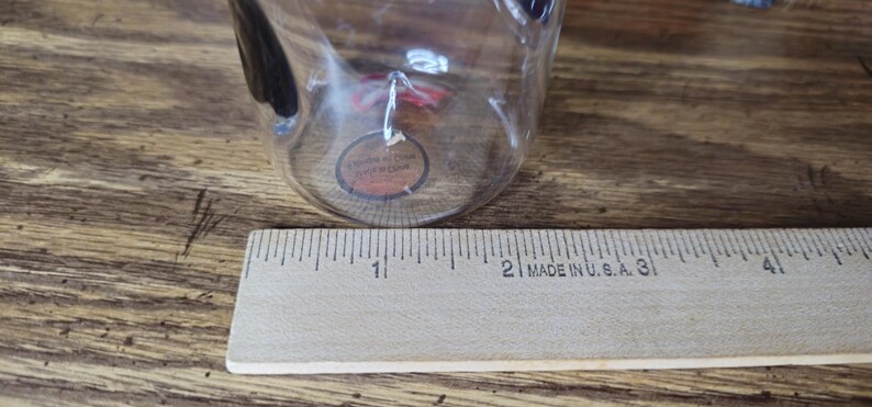 May include: A clear glass cup with a black handle is positioned near a wooden ruler. The ruler, marked in inches, is labeled "MADE IN U.S.A." The cup has a small, round sticker attached to it.