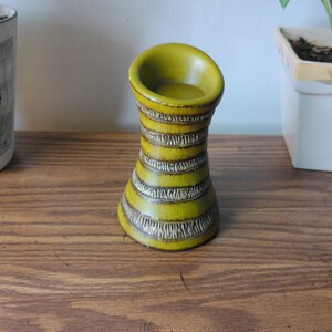 May include: A cylindrical ceramic candle holder featuring a vibrant olive green glaze with horizontal bands of textured, dark brown incised patterns. The object has a flared top opening and a tapered waist, showcasing a smooth finish on the rim.