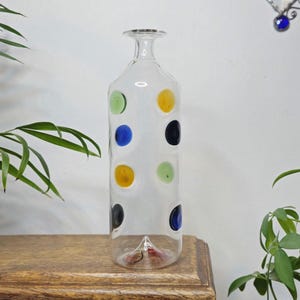 May include: Clear glass bottle with a narrow neck and a flat, rectangular body. The bottle is decorated with colorful polka dots in green, blue, yellow, and black. The bottle is displayed on a wooden surface.