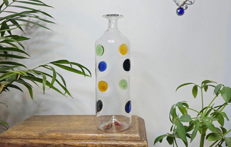 May include: Clear glass bottle with a narrow neck and a flat, rectangular body. The bottle is decorated with colorful polka dots in green, blue, yellow, and black. The bottle is displayed on a wooden surface.