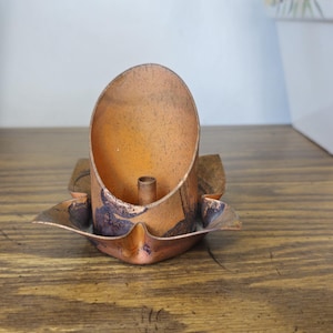 May include: A copper candle holder featuring a central cylindrical tube for a candle, surrounded by a wide, petal-like base with a weathered, dark patina finish. The metal surface shows signs of oxidation and texture throughout the structure.