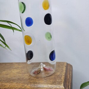 May include: A clear glass vase with colorful polka dots in green, blue, yellow, and black. The vase is cylindrical and sits on a wooden surface. Decorative vase for home decor.