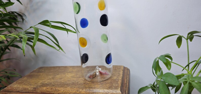 May include: A clear glass vase with colorful polka dots in green, blue, yellow, and black. The vase is cylindrical and sits on a wooden surface. Decorative vase for home decor.