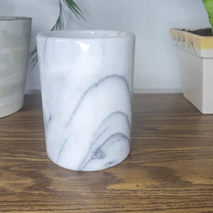 May include: A cylindrical white marble vessel featuring prominent grey veining throughout its smooth, polished surface. The container has a straight, vertical profile with a wide, open top and a flat base, showcasing natural stone patterns.