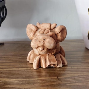May include: A small, tan-colored, handcrafted sculpture of a whimsical creature with large ears and a ruffled collar. The sculpture is made of a textured material and sits on a wooden surface. The creature has a smiling expression.