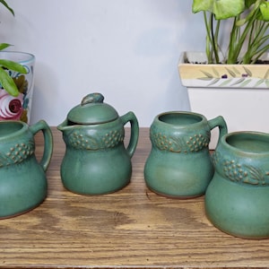 May include: A set of four teal ceramic mugs and a creamer with a lid, all with a textured grape vine design around the top. The mugs and creamer are arranged on a wooden surface, with plants in the background.