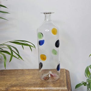 May include: A tall, clear glass bottle with a narrow neck and a flat base. The bottle is decorated with colorful polka dots in yellow, green, blue, and black. The bottle is displayed on a wooden surface.
