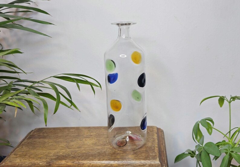 May include: A tall, clear glass bottle with a narrow neck and a flat base. The bottle is decorated with colorful polka dots in yellow, green, blue, and black. The bottle is displayed on a wooden surface.