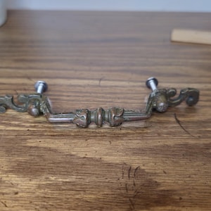 May include: An antique brass drawer pull with ornate detailing. The pull has a curved handle with decorative flourishes on each end and two screws for mounting. The hardware is set against a wooden surface.