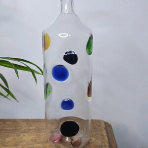 May include: A clear glass bottle with colorful polka dots in yellow, green, blue, black, and brown. The bottle is tall and cylindrical, with a narrow neck and a wide base. The bottle is sitting on a wooden surface.