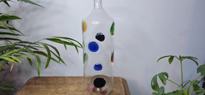 May include: A clear glass bottle with colorful polka dots in yellow, green, blue, black, and brown. The bottle is tall and cylindrical, with a narrow neck and a wide base. The bottle is sitting on a wooden surface.