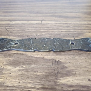 May include: A weathered, metal decorative hinge with a textured surface and two holes for mounting. The hinge has a unique, stylized design with decorative ends. The metal is a dark, aged color, suggesting an antique or vintage item.