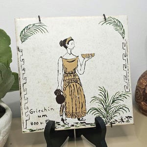 May include: A vintage tile depicting a woman in a yellow dress holding a bowl of fruit. The tile has a white background with green foliage and a Greek key border. The text "Griechin um 400 v." is printed on the tile.