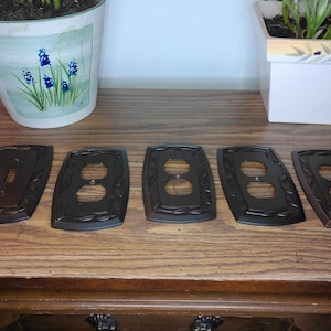 Op de afbeelding: Een set van vijf donkerbruine metalen wandplaten met een decoratieve bladrand. De collectie bevat één afdekplaat voor een tuimelschakelaar en vier afdekplaten voor dubbele stopcontacten, allemaal afgewerkt in een egale bronzen kleur.