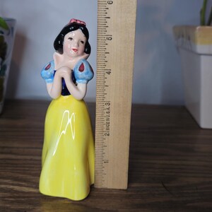 K&ouml;nnte beinhalten: Eine Keramikfigur von Schneewittchen mit gelbem Kleid, blauen und wei&szlig;en &Auml;rmeln und einer roten Schleife. Die Figur ist etwa 13 cm hoch, mit einem Lineal daneben zur Gr&ouml;&szlig;enbestimmung. Dekorative T&ouml;pfe sind im Hintergrund.