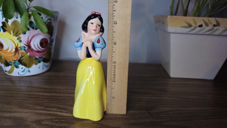 K&ouml;nnte beinhalten: Eine Keramikfigur von Schneewittchen mit gelbem Kleid, blauen und wei&szlig;en &Auml;rmeln und einer roten Schleife. Die Figur ist etwa 13 cm hoch, mit einem Lineal daneben zur Gr&ouml;&szlig;enbestimmung. Dekorative T&ouml;pfe sind im Hintergrund.