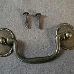 May include: Antique bronze-finished drawer pull with two matching screws. The pull has an oval backplate with a beaded edge and a curved handle. Ideal for furniture restoration or adding a vintage touch.