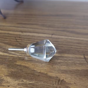 May include: A clear, faceted glass cabinet knob with a silver-colored stem. The knob is diamond-shaped and reflects light. The hardware is set against a wooden surface.