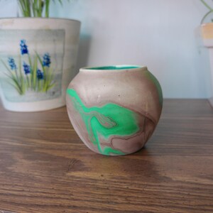 May include: A small, round ceramic vase featuring a marbled pattern of muted taupe and vibrant green swirls. The interior of the vase is finished in a solid, bright green glaze, while the exterior surface displays a smooth, matte texture.