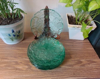 May include: A stack of green glass plates with a textured surface and a fruit design. The plates are stacked on a wooden surface. A brown decorative stand holds a clear glass plate with a fruit design. Two potted plants are in the background.