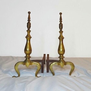 May include: Two antique brass and bronze fireplace andirons. Each andiron has a tall, ornate, vertical design with a rounded top and curved legs. A dark brown, wooden fire grate is between the andirons.