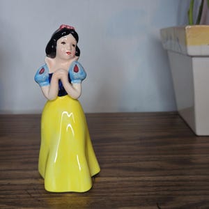 K&ouml;nnte beinhalten: Eine Keramikfigur von Schneewittchen mit gelbem Kleid, blauen und wei&szlig;en &Auml;rmeln und schwarzem Haar mit roter Schleife. Die Figur steht auf einer Holzoberfl&auml;che, mit einem Blumentopf und einem wei&szlig;en Pflanzgef&auml;&szlig; im Hintergrund.