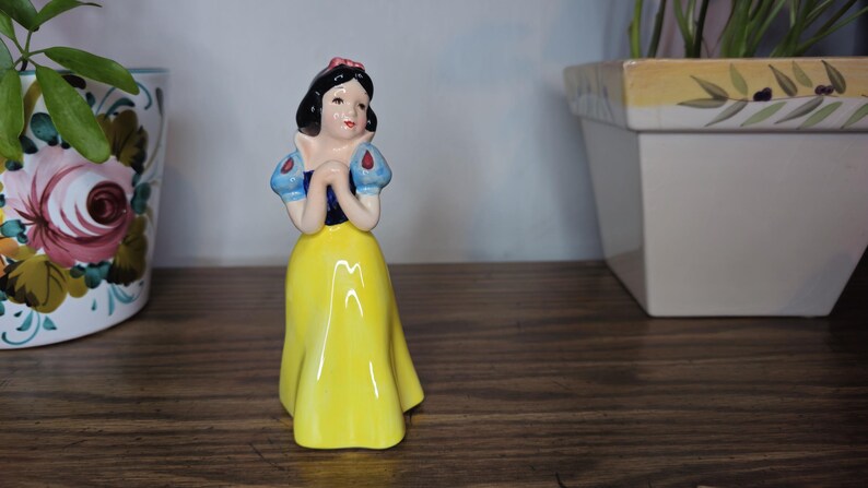 K&ouml;nnte beinhalten: Eine Keramikfigur von Schneewittchen mit gelbem Kleid, blauen und wei&szlig;en &Auml;rmeln und schwarzem Haar mit roter Schleife. Die Figur steht auf einer Holzoberfl&auml;che, mit einem Blumentopf und einem wei&szlig;en Pflanzgef&auml;&szlig; im Hintergrund.