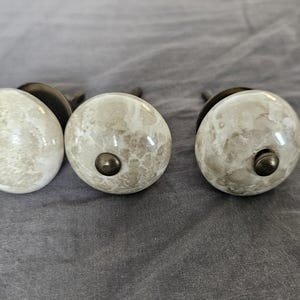 May include: Three marble-look cabinet knobs with a speckled, off-white and gray pattern. Each knob has a dark metal base and a small, dark metal screw. The knobs are round and sit on a gray surface.