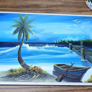 May include: A framed painting of a tropical beach scene. A palm tree leans over the shore, with a small boat and pier in the distance. The artwork features shades of blue and green, with white accents for the waves and sky. The artist's signature is visible.