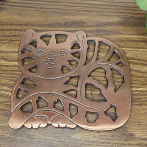 May include: Copper-colored metal cat-shaped trivet with cut-out details. The trivet features a stylized cat design with a smiling face, ears, and a curled tail. The cat measures approximately 6 inches long and 5 inches wide.