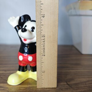 K&ouml;nnte beinhalten: Eine Mickey Mouse-Figur, etwa 7,6 cm hoch, in einer klassischen Pose mit einem erhobenen Arm. Die Figur ist schwarz, rot, wei&szlig; und gelb. Ein Holzlineal befindet sich neben der Figur.