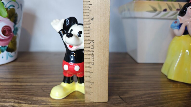 K&ouml;nnte beinhalten: Eine Mickey Mouse-Figur, etwa 7,6 cm hoch, in einer klassischen Pose mit einem erhobenen Arm. Die Figur ist schwarz, rot, wei&szlig; und gelb. Ein Holzlineal befindet sich neben der Figur.