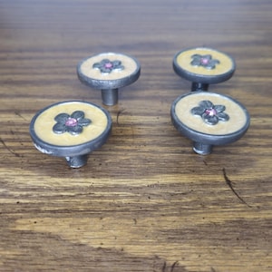 May include: Four round cabinet knobs with a dark metallic finish and a light beige circular inlay. Each knob features a dark gray flower-shaped metal embellishment in the center with a small, singular pink rhinestone embedded in the middle.