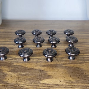 May include: A collection of eleven dark metallic cabinet knobs arranged in rows. Each knob features a circular, slightly domed top with a smooth, reflective finish and a tapered base that connects to a central mounting point for furniture attachment.