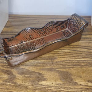 May include: A rectangular ceramic tray with a metallic bronze glaze and a wavy, undulating rim. The interior surface features a textured pattern of small dots and geometric lines, while the ends are finished with rolled, scroll-like handles.