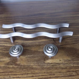 May include: Two silver-toned metal cabinet pulls with a horizontal wavy design and two matching silver-toned metal cabinet knobs featuring a circular spiral pattern. These hardware pieces have a smooth, matte finish and are designed for furniture.