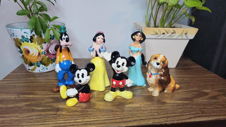 K&ouml;nnte beinhalten: Eine Sammlung von Keramikfiguren mit Disney-Charakteren, darunter Micky Maus, Schneewittchen, Jasmin und Goofy. Die Figuren sind farbenfroh und auf einer Holzoberfl&auml;che angeordnet, die ein nostalgisches Thema zeigt.