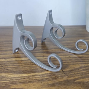 May include: A pair of silver-colored metal wall hooks featuring a curved, scroll-like design. Each hook has a vertical mounting plate with two pre-drilled screw holes at the top and bottom for secure attachment to a flat surface.