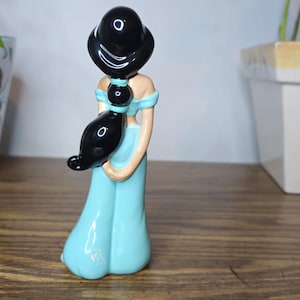 K&ouml;nnte beinhalten: Keramikfigur einer Frau in einem hellblauen Kleid mit schwarzen Haaren. Die Figur hat ein detailliertes Design und steht auf einer Holzoberfl&auml;che. Die Figur ist ein Sammlerst&uuml;ck.