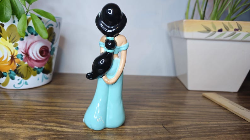 K&ouml;nnte beinhalten: Keramikfigur einer Frau in einem hellblauen Kleid mit schwarzen Haaren. Die Figur hat ein detailliertes Design und steht auf einer Holzoberfl&auml;che. Die Figur ist ein Sammlerst&uuml;ck.