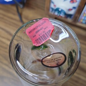 May include: Clear glass vase with a pink tag that reads "ANTHROPOLOGIE". The vase has a round base and a cylindrical shape. A sticker on the bottom says "Made in China".