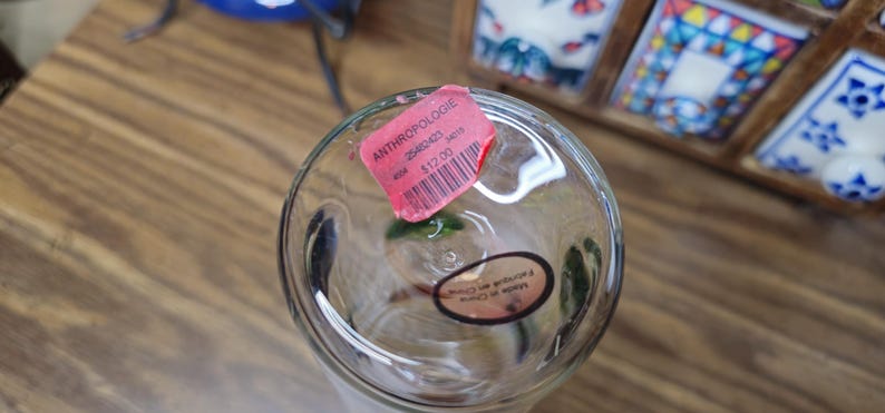 May include: Clear glass vase with a pink tag that reads "ANTHROPOLOGIE". The vase has a round base and a cylindrical shape. A sticker on the bottom says "Made in China".