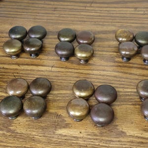 May include: A collection of antique brass cabinet knobs in various shades of bronze and gold. The knobs are arranged in groups on a wooden surface, showcasing their aged patina and classic design. Ideal for furniture restoration.