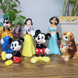K&ouml;nnte beinhalten: Eine Sammlung von Keramikfiguren mit Disney-Charakteren, darunter Micky Maus, Schneewittchen, Jasmin, Goofy und ein Hund. Die Figuren sind farbenfroh und auf einer Holzoberfl&auml;che angeordnet.