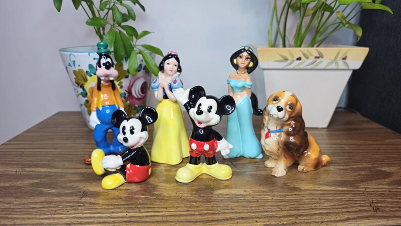 K&ouml;nnte beinhalten: Eine Sammlung von Keramikfiguren mit Disney-Charakteren, darunter Micky Maus, Schneewittchen, Jasmin, Goofy und ein Hund. Die Figuren sind farbenfroh und auf einer Holzoberfl&auml;che angeordnet.