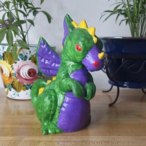 May include: A whimsical ceramic dragon figurine in shades of green and purple, with yellow accents. The dragon has red eyes and is posed on a wooden surface. Decorative item.
